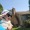 baby, blurred_background, building, casual, child, daylight, grass, greenery, hand, happy, hat, holding, outdoor, person, portrait, skin, smiling, sunglasses, sunny, white_clothing