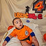 baby, backdrop, birthday_decoration, blanket, blue_socks, blue_wristband, child, cute, decorative_letters, dragon_ball, gaze, goku_silhouette, infant, number_4, orange_outfit, plush, portrait, sitting, studio_lighting, watermark