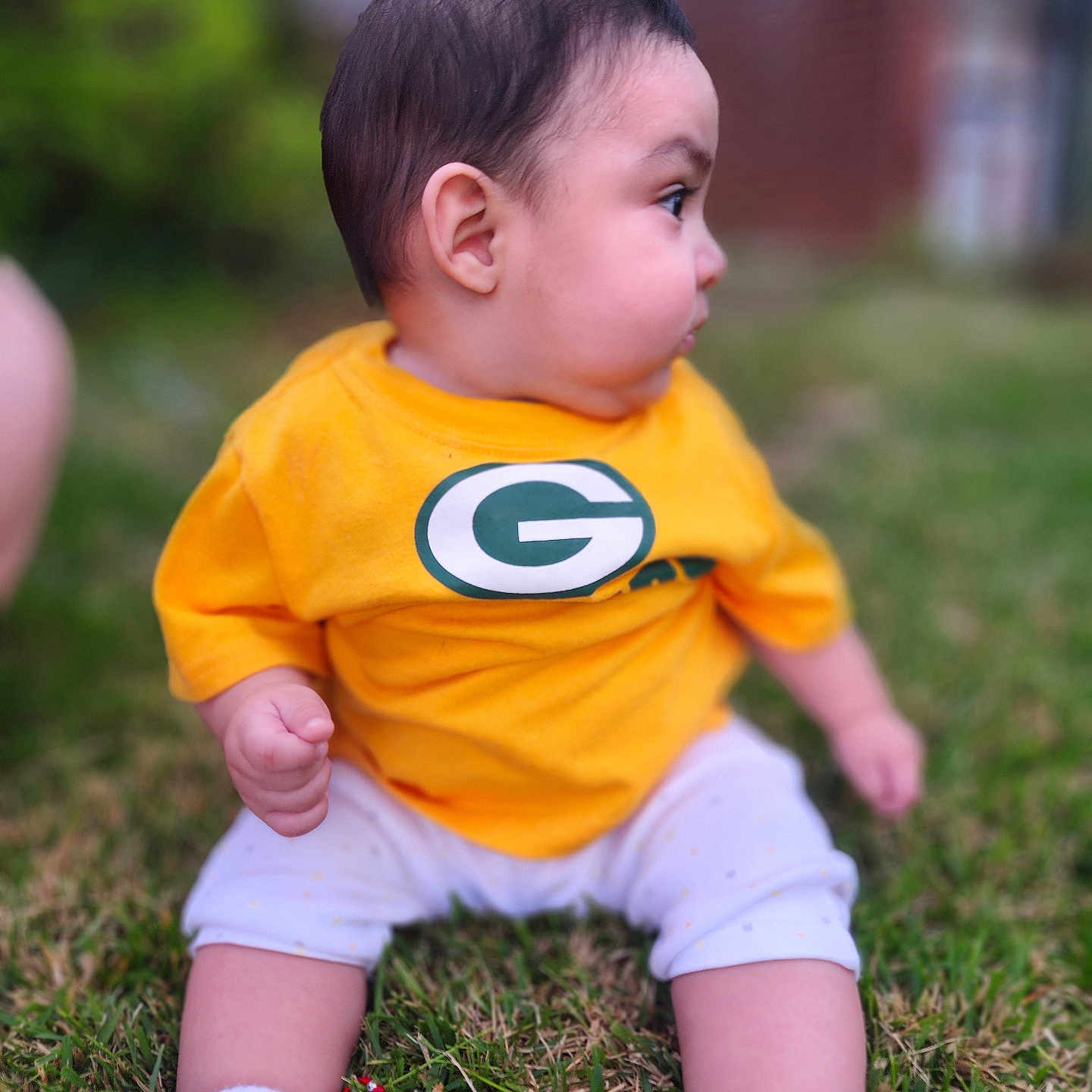 Bodoquito is registered to the contest to win money with this photo: baby, child, grass, yellow_shirt, socks, outdoor, side_profile, curious, sitting, green_logo, short_hair, infant, casual_clothing, nature, daylight, cute, young_child, portrait, small_hands, feet