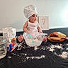 apron, baby, baking, chef_hat, child, cookies, cute, doughnut, flour, food, indoor, kitchen, messy, mixing_bowl, pastries, person, playful, sitting, spoon, table
