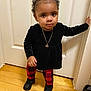 black_bow, black_sneakers, black_top, child, curly_hair, cute, door, fashion, floor, hand_on_door, indoors, necklace, person, portrait, red_checkered_leggings, standing, toddler, velvet, wooden_floor, young_child