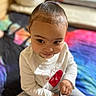 toddler, child, face, smile, white_shirt, heart_design, sitting, indoors, bed, colorful_bedspread, toy, footwear, curly_hair, cute, expression, playful, person, portrait, young_child, casual_clothing