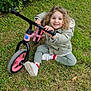 Gabriela participe au concours pour gagner de l'argent avec cette photo : balance_bike, casual_clothing, child, curly_hair, daylight, fun, grass, greenery, happy, jacket, nature, outdoor, park, pink_bike, playful, shoes, sitting, smiling, toddler, young_child