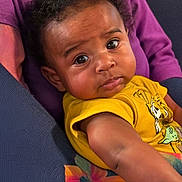 Cortez is registered to the contest to win money with this photo: baby, child, curly_hair, yellow_shirt, cartoon_print, floral_pattern, purple_cloth, face, portrait, indoors, person, cute, expression, skin, arm, seated, closeup, young, infant, looking