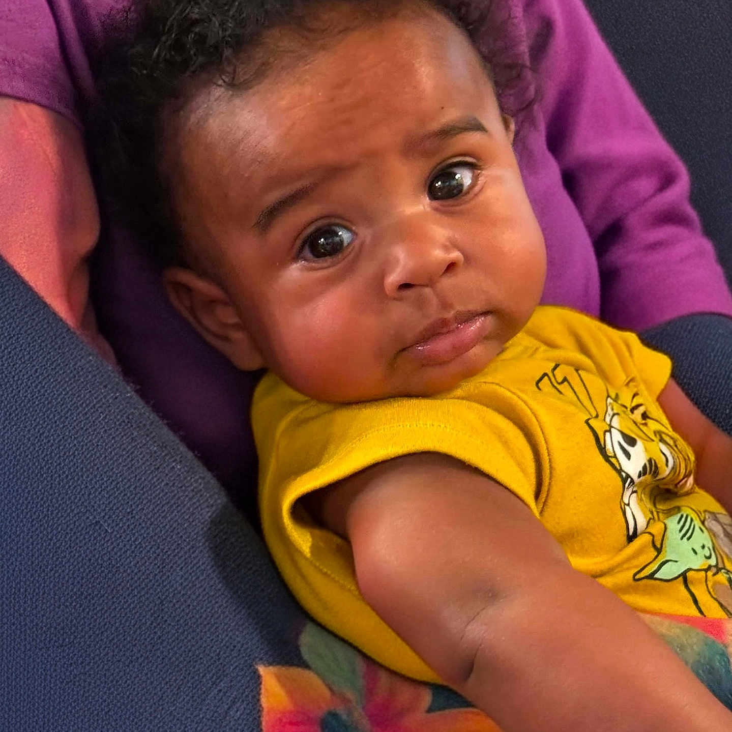 Cortez is registered to the contest to win money with this photo: baby, child, curly_hair, yellow_shirt, cartoon_print, floral_pattern, purple_cloth, face, portrait, indoors, person, cute, expression, skin, arm, seated, closeup, young, infant, looking