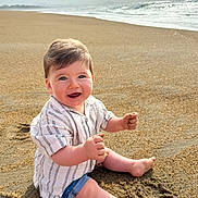 éli a rejoint le concours — aidez-le/la à gagner de superbes lots ! baby, child, sand, beach, ocean, water, waves, smiling, happy, sitting, clothing, shorts, shirt, barefoot, sunlight, sky, clouds, nature, outdoor, portrait