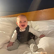 Alys joined the competition — help win amazing prizes! baby, smiling, bed, pillow, white_bedding, wooden_headboard, infant, happy, child, indoor, cute, sitting, portrait, cozy, soft_light, clothing, sock, person, cheerful, young_child