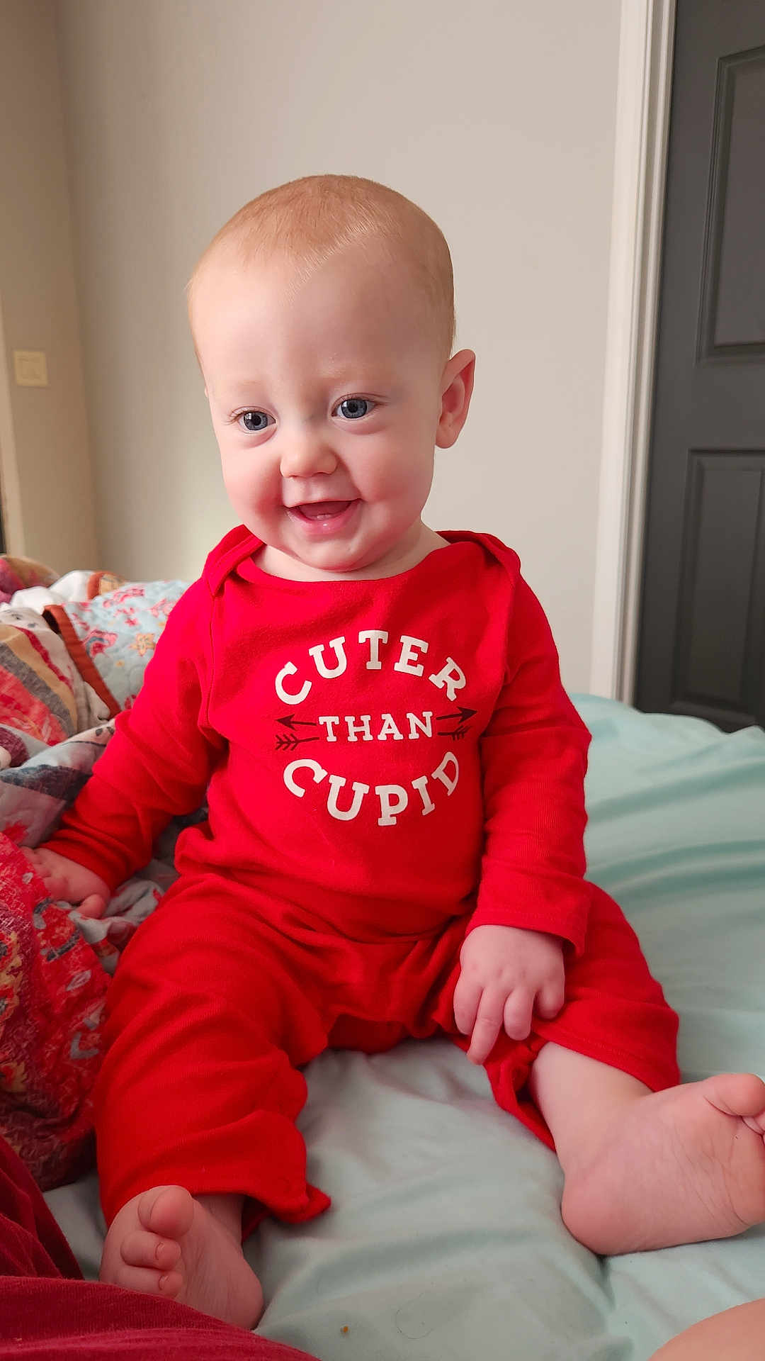 Parker joined the competition — help win amazing prizes! baby, child, red_clothing, onesie, smile, happy, indoors, bed, blanket, foot, hand, face, cute, sitting, infant, person, skin, room, door, text_print