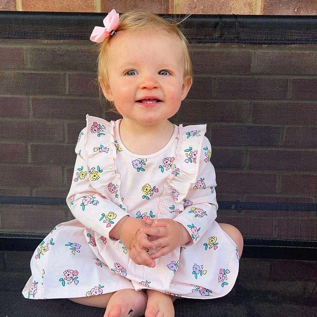 Darcy Leigh is registered to the contest to win money with this photo: baby, barefoot, bench, brick_wall, child, cute, daylight, dress, floral_pattern, girl, hands_clasped, happy, mesh_bench, outdoor, person, pink_bow, portrait, sitting, smiling, toddler