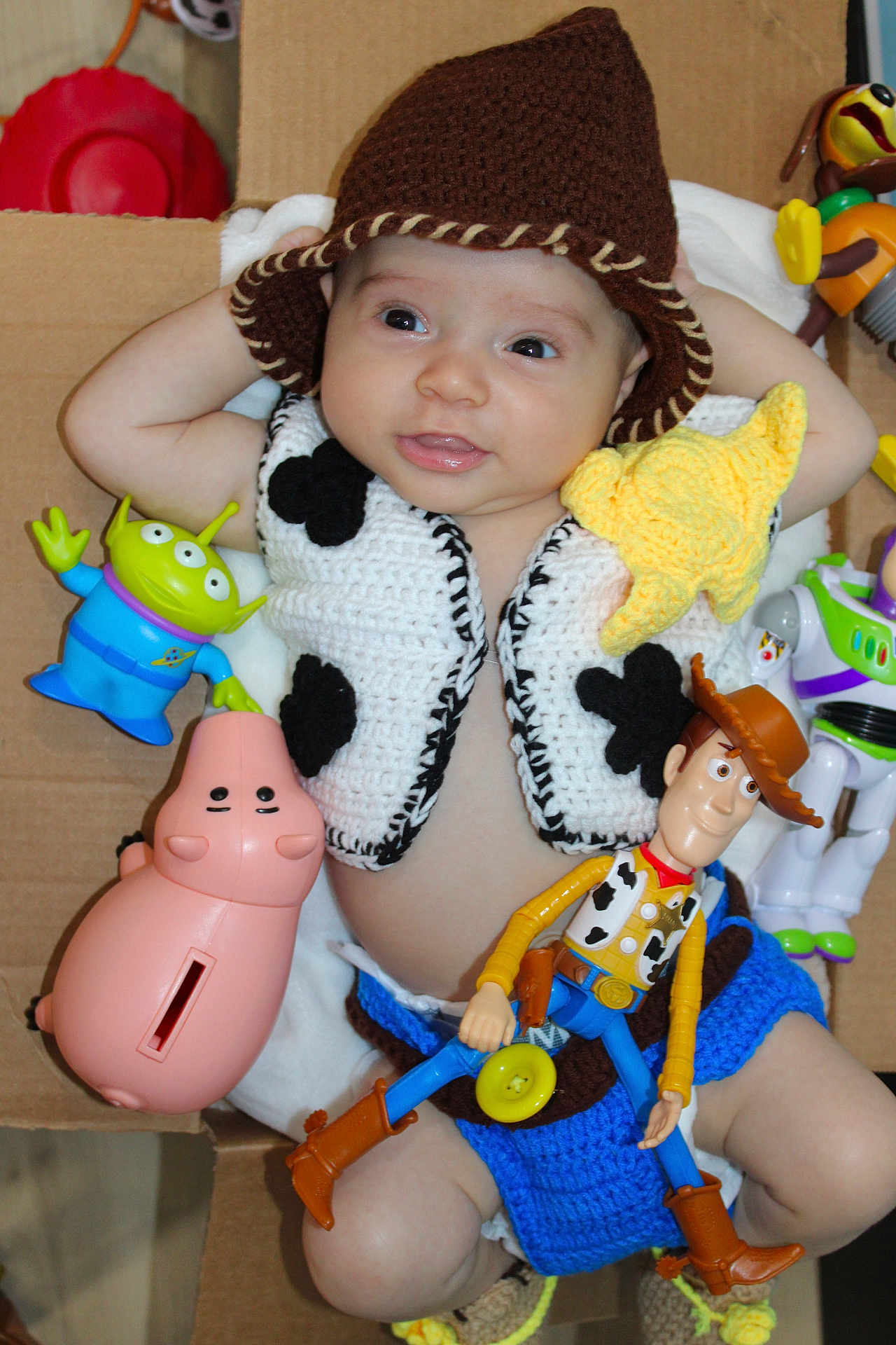 Thiago is registered to the contest to win money with this photo: baby, child, cowboy, crocheted_clothing, hat, toy_story, woody, buzz_lightyear, alien_toy, pink_piggy_bank, yellow_star, cardboard_box, cute, smiling, indoor, portrait, toy, infant, costume, playful