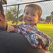 Waylan joined the competition — help win amazing prizes! baby, child, smiling, blanket, cartoon_pattern, outdoor, fence, sports_field, adult, shoulder, happy, cute, daylight, person, portrait, grass, head, clothing, sky, playful