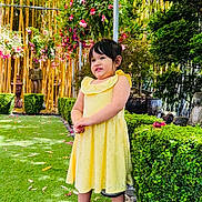 Angelina is registered to the contest to win money with this photo: child, girl, yellow_dress, green_grass, flowers, bamboo, garden, plants, hedge, statue, outdoor, smiling, person, nature, shoes, sunlight, daytime, happy, portrait, standing