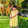 child, girl, yellow_dress, green_grass, flowers, bamboo, garden, plants, hedge, statue, outdoor, smiling, person, nature, shoes, sunlight, daytime, happy, portrait, standing