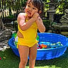 barefoot, blue_pool, child, flowers, garden, grass, greenery, happy, nature, outdoor, person, playful, portrait, stone_sculpture, summer, sunlight, toddler, toys, water_play, yellow_swimsuit