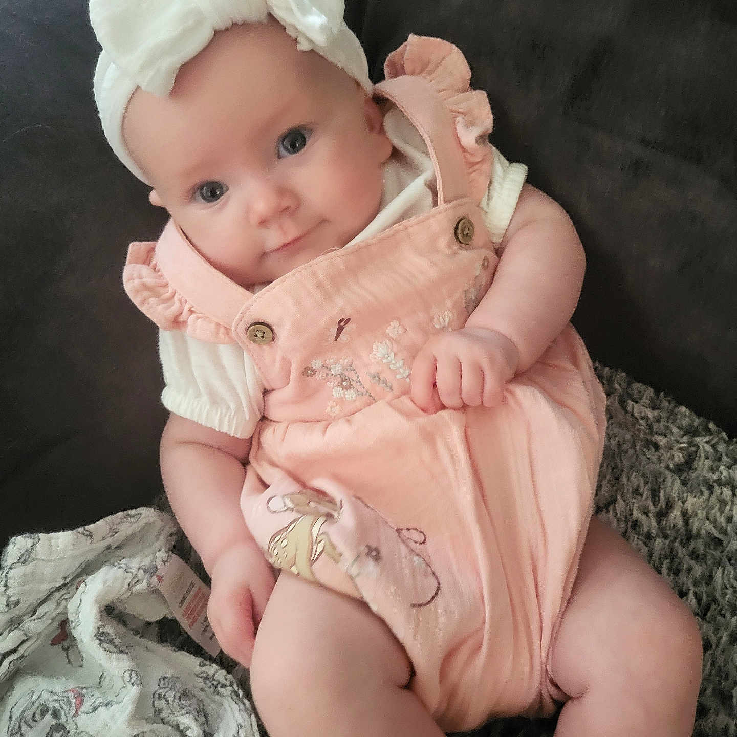 Lyra-Lily is registered to the contest to win money with this photo: baby, blanket, bow, cartoon_print, child, chubby_cheeks, closeup, couch, curious, cute, headband, indoor, infant, person, pink_clothing, portrait, romper, seated, smiling, soft_lighting