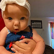 Kailani is registered to the contest to win money with this photo: baby, infant, child, headband, white_bow, blue_dress, hand, adult_hand, indoor, living_room, curtains, air_conditioner, skin, person, cute, portrait, expression, holding, furniture, table