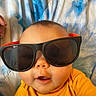 Kailani is registered to the contest to win money with this photo: baby, child, sunglasses, yellow_shirt, face, portrait, cute, infant, lying_down, blanket, blue, orange, skin, head, eyes, mouth, hand, clothing, indoor, closeup
