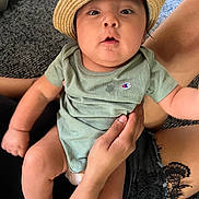 Mateo is registered to the contest to win money with this photo: baby, carpet, child, closeup, clothing, curious, cute, face, hand, hat, holding, indoor, infant, legs, onesie, person, sitting, skin, surprised, tattoo