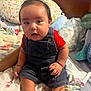 Mateo is registered to the contest to win money with this photo: baby, bed, child, curious, cute, denim_overalls, face, floral_pattern, footwear, hands, head, home, indoor, infant, person, pillow, portrait, red_shirt, sitting, skin