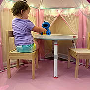 Amelia is registered to the contest to win money with this photo: toddler, child, toy, cookie_monster, table, chair, play_tent, pink, purple_shirt, indoor, wooden_chair, floor_mat, curtains, string_lights, playroom, furniture, cute, small_table, childrens_room, playtime
