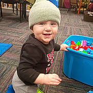 Landon is registered to the contest to win money with this photo: toddler, child, indoor, playing, toys, blocks, knit_hat, carpet, sweatshirt, smiling, floor, colorful, blue_container, furniture, person, happy, young_child, casual_clothing, development, kid
