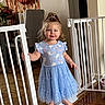 baby_gate, balloons, barefoot, blue_dress, chair, child, cute, festive, flowers, hair_tied, happy, home, indoor, person, portrait, smiling, standing, table, toddler, wooden_floor