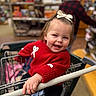 Hadley Dawn is registered to the contest to win money with this photo: toddler, child, shopping_cart, red_sweater, bow, smiling, happy, indoors, person, blurred_background, shopping, clothing, hand, face, hair, store, floor, footwear, sitting, cute