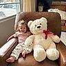 barefoot, casual_clothing, chair, child, curtain, cushion, happy, home_interior, indoor, leather, living_room, natural_light, plant, plush_toy, red_bow, relaxed, smiling, teddy_bear, toddler, window
