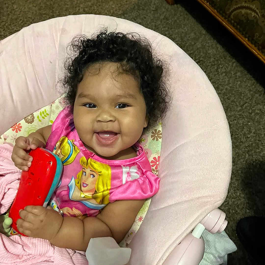 Halo Smith is registered to the contest to win money with this photo: baby, baby_seat, carpet, child, colorful, comfort, cozy, curly_hair, cute, face, happy, indoor, infant, person, pink_dress, playful, portrait, seated, smiling, toy