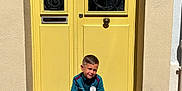 Antoine a rejoint le concours — aidez-le/la à gagner de superbes lots ! child, boy, sitting, door, yellow_door, outdoor, sunlight, sportswear, shoes, socks, shorts, wall, building, street, portrait, casual, smile, hair, concrete, urban