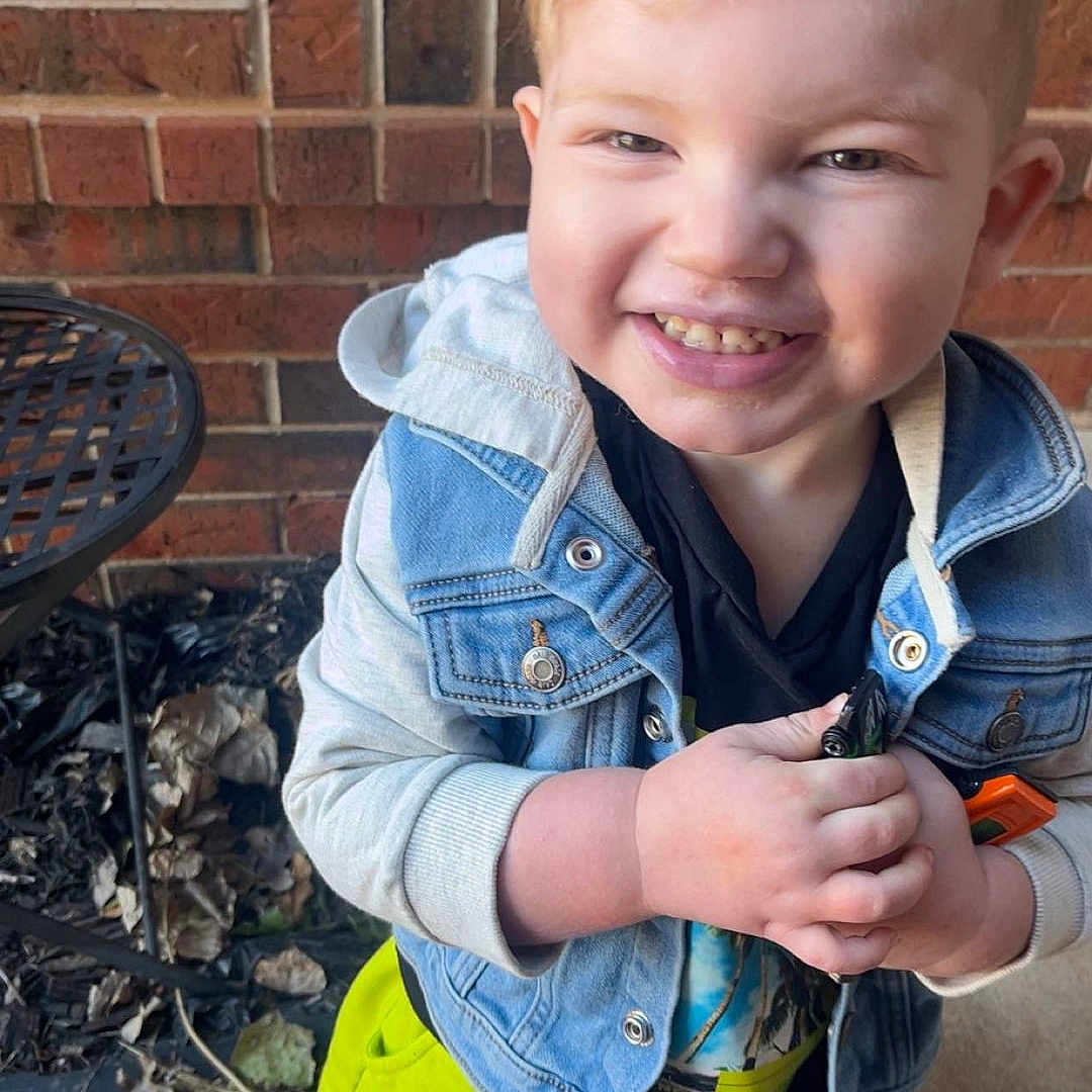 Graylen joined the competition — help win amazing prizes! blonde_hair, brick_wall, casual_clothing, child, concrete, cute, daylight, denim_jacket, expression, happy, holding, metal_chair, outdoor, person, portrait, smiling, standing, toddler, white_shoes, yellow_shorts