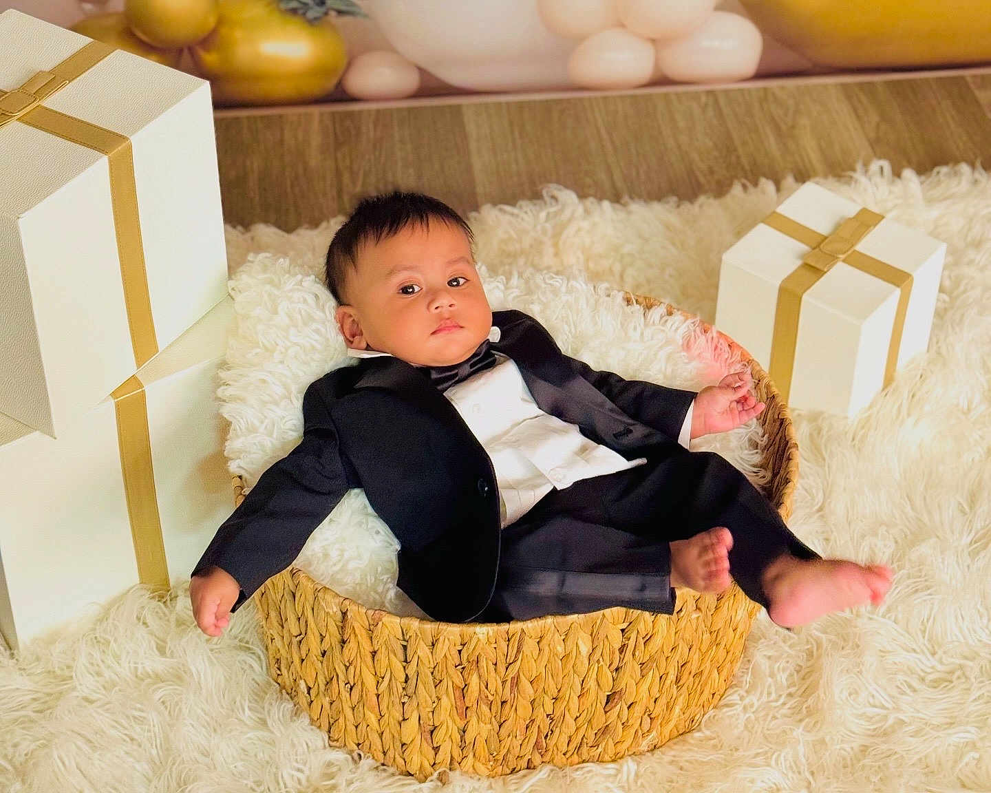 Ivaan joined the competition — help win amazing prizes! baby, tuxedo, basket, gift_box, white_rug, flooring, indoors, child, portrait, clothing, person, cute, formal_wear, infant, furniture, smile, celebration, holiday, decor, relaxing