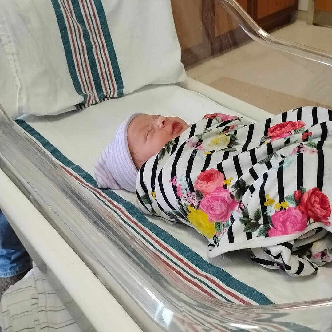 Destinee is registered to the contest to win money with this photo: newborn, baby, hospital, bassinet, blanket, striped, floral, sleeping, towel, hat, person, adult, hospital_room, cabinet, floor, medical_equipment, comfort, indoor, rest, care