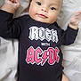 arms, baby, bed, bedding, black_clothing, child, cute, face, happy, indoor, infant, legs, lying_down, onesie, person, skin, smile, soft_light, text_on_clothing, young