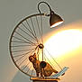 cat, tabby_cat, bicycle_wheel, lamp, light, shadow, wall, curious, indoor, feline, pet, animal, metal, cord, wooden_surface, decor, artistic, lighting, household, exploration
