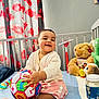 baby, ball, bed, cactus_toy, cardigan, child, colorful, curtain, floral_pattern, happy, indoor, nursery, person, photography, play, skirt, smiling, stuffed_animal, teddy_bear, toy
