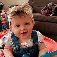 Shiloh is registered to the contest to win money with this photo: baby, child, smiling, headband, blue_eyes, denim_overalls, toy, dumbbell_toy, activity_center, blanket, living_room, couch, indoor, person, cute, happy, infant, playtime, pink_blanket, colorful_toys