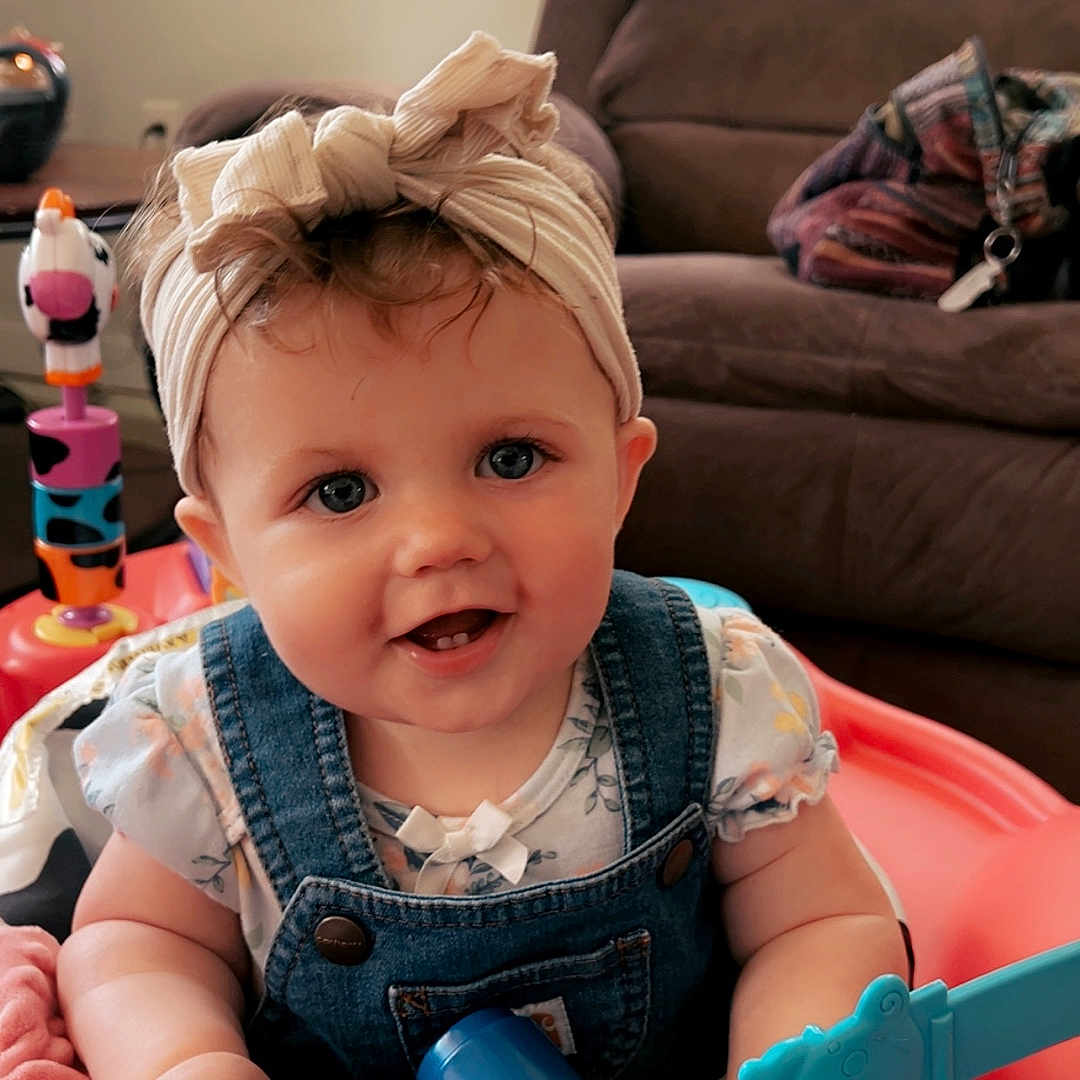 Shiloh is registered to the contest to win money with this photo: activity_center, baby, blanket, blue_eyes, child, colorful_toys, couch, cute, denim_overalls, dumbbell_toy, happy, headband, indoor, infant, living_room, person, pink_blanket, playtime, smiling, toy