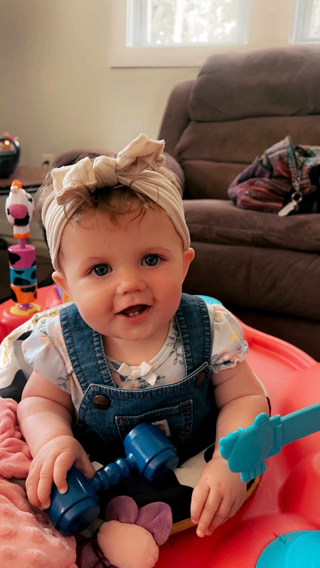 Shiloh is registered to the contest to win money with this photo: baby, child, smiling, headband, blue_eyes, denim_overalls, toy, dumbbell_toy, activity_center, blanket, living_room, couch, indoor, person, cute, happy, infant, playtime, pink_blanket, colorful_toys