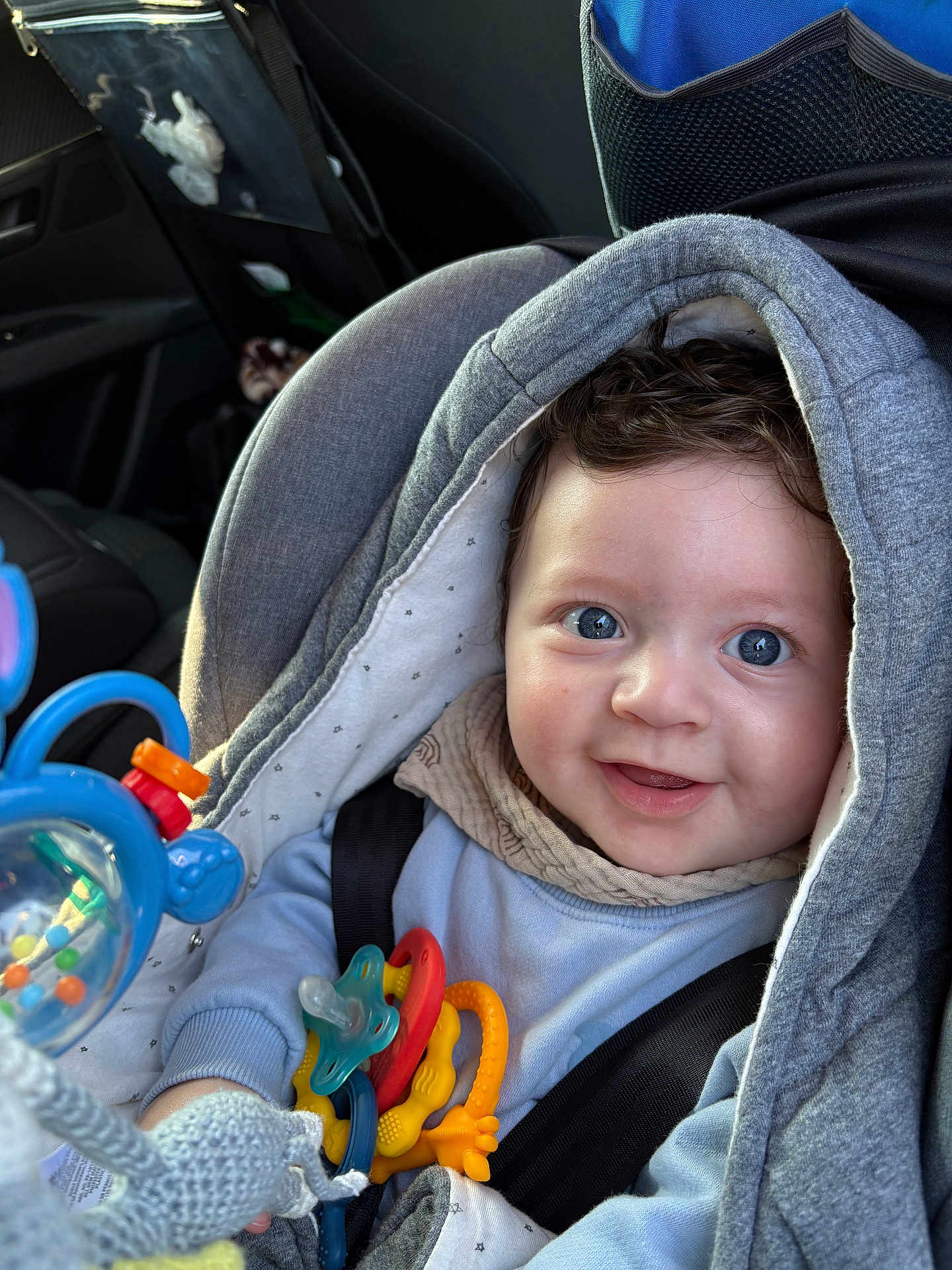 Rafaël a rejoint le concours — aidez-le/la à gagner de superbes lots ! baby, blue_eyes, car_seat, close_up, colorful, cozy, curly_hair, cute, fabric, hand, hooded_jacket, infant, pacifier, playtime, portrait, seatbelt, smiling, teether, toy, transport