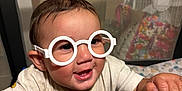 Legend is registered to the contest to win money with this photo: baby, child, glasses, playpen, toy, smile, happy, face, hand, clothing, indoor, pattern, animal_print, furniture, person, cute, infant, play, young, expression