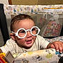 baby, child, glasses, playpen, toy, smile, happy, face, hand, clothing, indoor, pattern, animal_print, furniture, person, cute, infant, play, young, expression