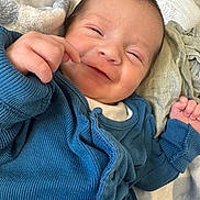 Reggie is registered to the contest to win money with this photo: baby, infant, smile, blue_clothing, cozy, blanket, hand, face, portrait, newborn, cute, happy, lying_down, soft_texture, closeup, skin, person, baby_clothes, warm, indoor