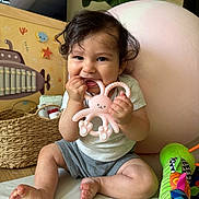 Ezra is registered to the contest to win money with this photo: baby, child, toy, teether, smiling, curly_hair, indoor, basket, playtime, cute, happy, colorful, soft_surface, couch, infant, person, chewing, pink, green, decor