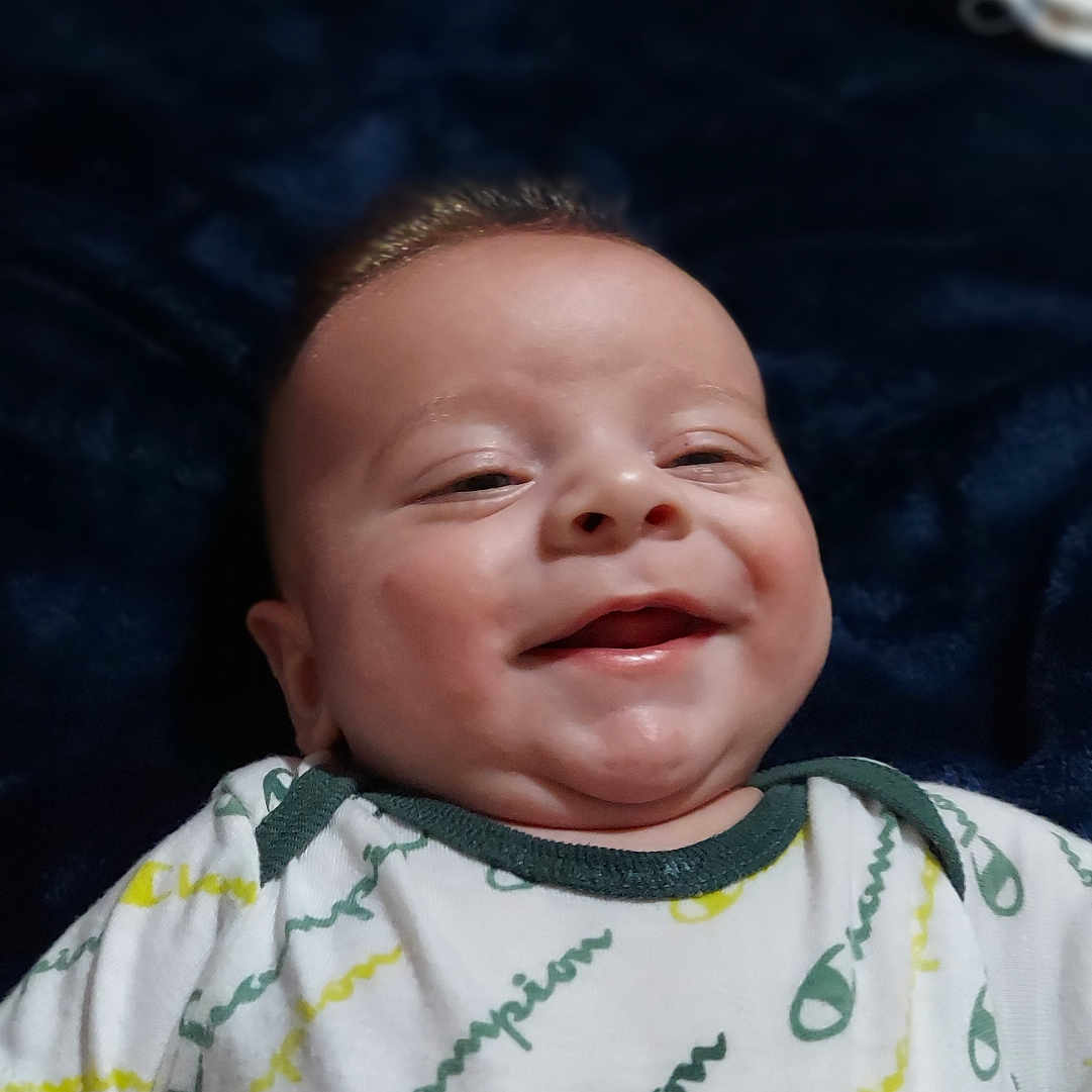 River Dudley is registered to the contest to win money with this photo: baby, smiling, infant, onesie, clothing, champion, blanket, portrait, happy, face, child, cute, lying_down, baby_garment, young_child, indoors, soft_lighting, skin, expression, cozy