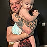 Vincent is registered to the contest to win money with this photo: arm, baby, black_shirt, book, child, colorful, curious, diaper, facial_hair, furniture, holding, indoor, long_hair, love, man, parent, person, room, skin, tattoo