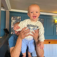Reuben is registered to the contest to win money with this photo: baby, child, infant, smiling, happy, portrait, indoor, adult, parent, holding, family, blue_wall, wooden_table, floral_panel, lamp, clothing, sweater, brooklyn_shirt, light_blue_pants, barefoot
