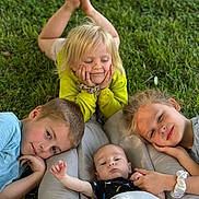 Nolan is registered to the contest to win money with this photo: baby, blanket, blonde_hair, brown_hair, casual_clothing, children, daylight, family, garden, grass, group_portrait, infant, lying_down, outdoor, playful, portrait, siblings, siblings_together, smiling, toddler