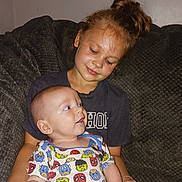 Nolan is registered to the contest to win money with this photo: child, baby, sibling, sister, infant, sleeping, awake, couch, sitting, portrait, indoors, family, 抱抱, smiling, brown_hair, bun, dark_clothing, superhero_print, blanket, cuddling