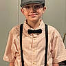 child, boy, smile, glasses, hat, cap, bow_tie, suspenders, shirt, portrait, indoor, person, fashion, casual_clothing, young, face, eyewear, standing, childhood, happy